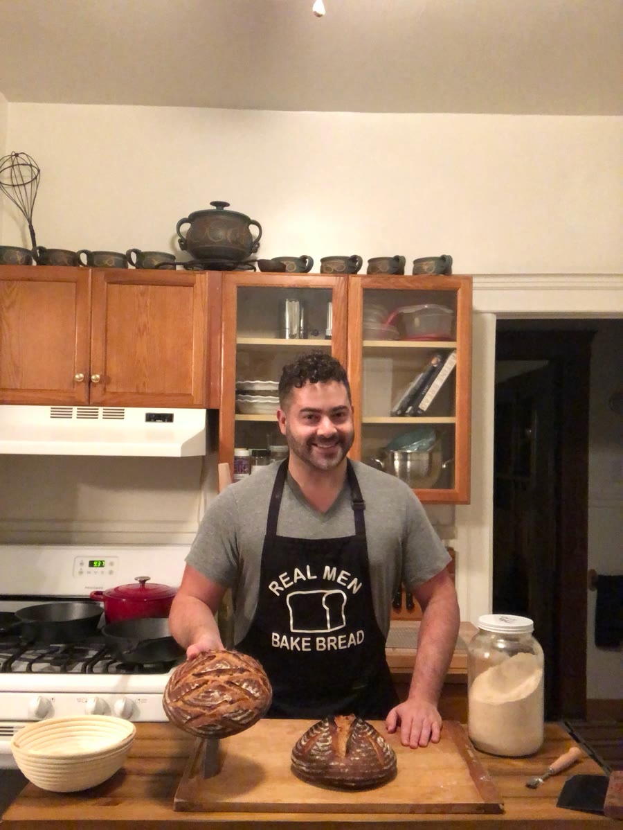 Arthur in his kitchen in a Real Men Bake Bread apron, holding two scored sourdough loaves